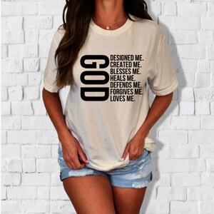 Pode incluir: Camiseta branca com texto preto que diz "GOD" verticalmente e "DESIGNED ME. CREATED ME. BLESSES ME. HEALS ME. DEFENDS ME. FORGIVES ME. LOVES ME." abaixo.
