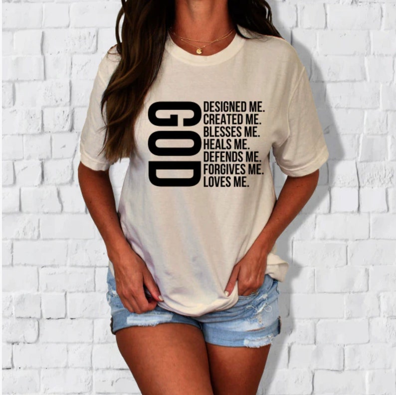Pode incluir: Camiseta branca com texto preto que diz "GOD" verticalmente e "DESIGNED ME. CREATED ME. BLESSES ME. HEALS ME. DEFENDS ME. FORGIVES ME. LOVES ME." abaixo.