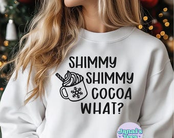May include: White sweatshirt with black text that reads "Shimmy Shimmy Cocoa What?" and a graphic of a cup of hot cocoa with whipped cream and a snowflake. The text "Janai's Tees" is in a smaller font below the graphic.