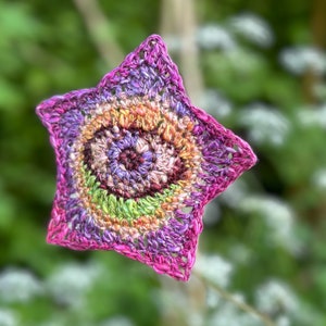 May include: A crocheted star with a multi-coloured spiral design in shades of pink, purple, green, and orange. The star is made with a fine yarn and has a delicate, lacy appearance.