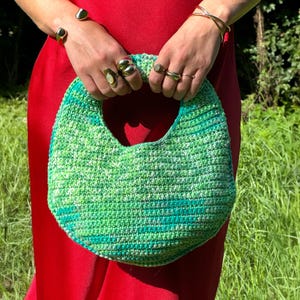 Op de afbeelding: Een groen en blauw gehaakte handtas met een afgeronde vorm en een grote handgreepopening. De tas wordt tegen een rode achtergrond gehouden. De textuur van de tas is zichtbaar. De handen van een persoon die de tas vasthoudt, zijn zichtbaar.