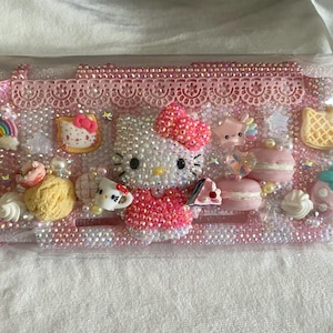 May include: A pink handheld gaming console case decorated with numerous small beads, a central Hello Kitty figure, and various miniature charms including a rainbow, macarons, a waffle, and ice cream, all encased in a clear plastic protective sleeve.