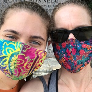 May include: Two people wearing face masks. The person on the left is wearing a colorful patterned mask with yellow, blue, pink, and green colors. The person on the right is wearing a black floral patterned mask with red, pink, and blue flowers.