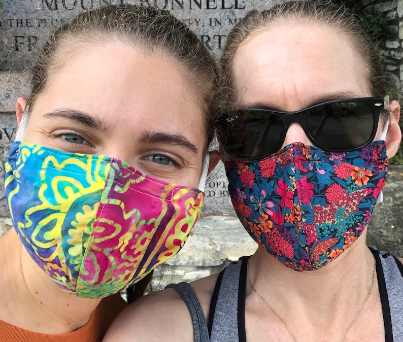 May include: Two people wearing face masks. The person on the left is wearing a colorful patterned mask with yellow, blue, pink, and green colors. The person on the right is wearing a black floral patterned mask with red, pink, and blue flowers.