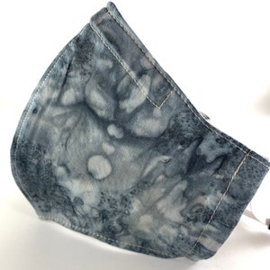 May include: A gray and white tie-dye fabric face mask with white stitching. The mask is made of a soft, breathable material and has a comfortable fit.