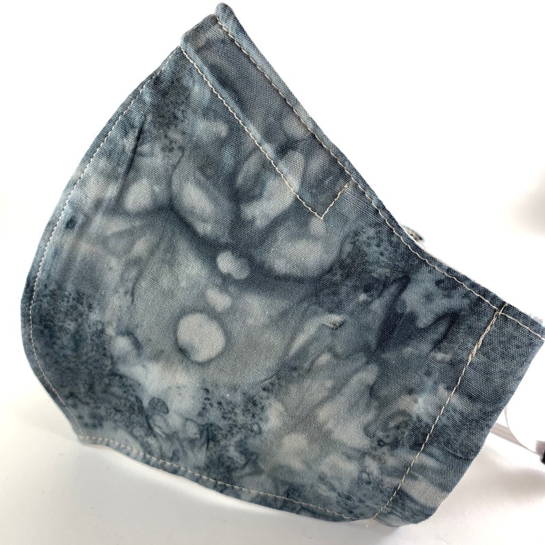 May include: A gray and white tie-dye fabric face mask with white stitching. The mask is made of a soft, breathable material and has a comfortable fit.