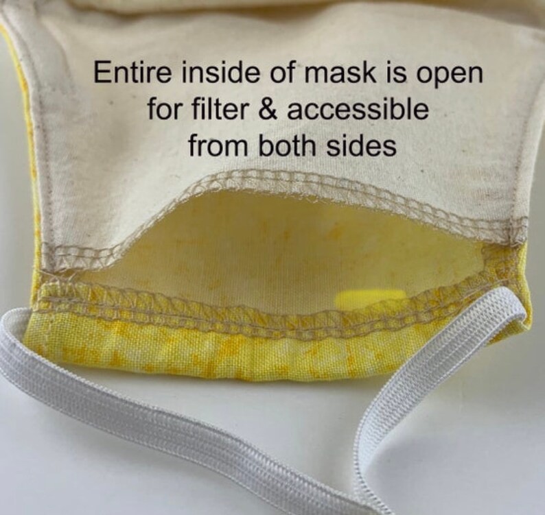 May include: A yellow fabric face mask with a white elastic band. The inside of the mask is open for a filter and is accessible from both sides. The text "Entire inside of mask is open for filter & accessible from both sides" is printed on the mask.