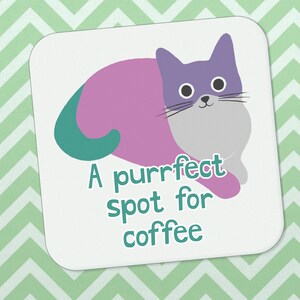 May include: A white square coaster featuring a purple and teal cat curled up. The text "A purrfect spot for coffee" is written in teal below the cat.