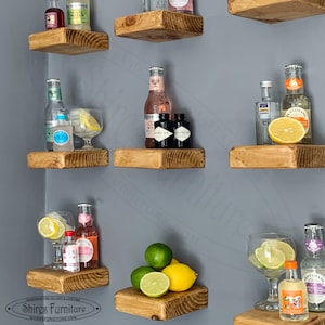 May include: A set of wooden shelves with various bottles of spirits, mixers, and garnishes. The shelves are stained a light brown colour and have a rustic appearance. The bottles are arranged in a decorative manner, with some bottles placed on top of others. The shelves are mounted on a grey wall.