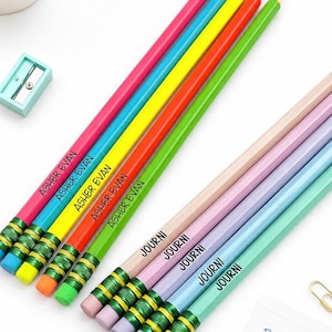 May include: A set of ten wooden pencils with erasers, featuring five bright colors labeled ASHER EVAN and five pastel colors labeled JOURNI. The pencils are arranged diagonally alongside a light blue pencil sharpener and a note reading Ready for school!