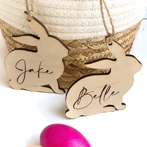 May include: Two wooden bunny shaped Easter tags with the names Jake and Belle written on them. The tags are hanging from a woven basket. Two Easter eggs, one pink and one yellow, are in the foreground.