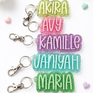 May include: Five rectangular resin keychains with scalloped edges and silver-tone lobster clasps. Each keychain features glitter and a raised name in a contrasting color: AKIRA in yellow, AVY in pink, KAMILLE in purple, JANIYAH in teal, and MARIA in green.