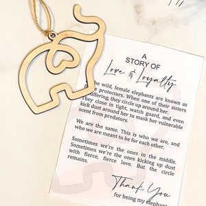 May include: A wooden elephant ornament with a pink and white background card that reads "A Story of Love & Loyalty". The card has a quote about female elephants protecting each other and ends with "Thank You for being my elephant".