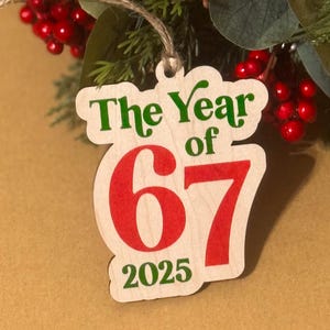 May include: Wooden Christmas ornament with the text "The Year of 67 2025" in green and red. The ornament is suspended by a twine string and is surrounded by greenery and red berries.