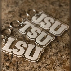 Könnte beinhalten: Drei transparente Acryl-Schlüsselanhänger mit den Buchstaben "LSU" in Weiß. Die Schlüsselanhänger sind übereinander gestapelt.