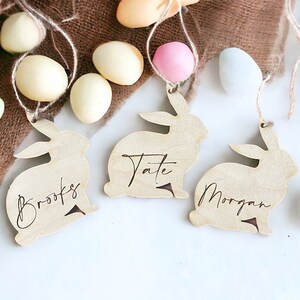 May include: Three wooden bunny-shaped ornaments with the names Brooks, Tate, and Morgan written on them. The ornaments are hanging from twine and are surrounded by pastel-colored Easter eggs.