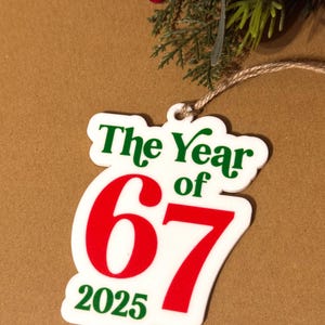 K&ouml;nnte beinhalten: Ein wei&szlig;es Ornament mit dem Text "The Year of 67 2025" in Gr&uuml;n und Rot. Das Ornament h&auml;ngt an einer Schnur, mit roten Beeren und gr&uuml;nem Laub im Hintergrund. Das Ornament ist wahrscheinlich f&uuml;r einen Feiertag oder einen besonderen Anlass.