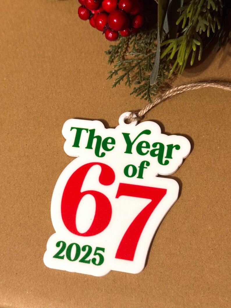 K&ouml;nnte beinhalten: Ein wei&szlig;es Ornament mit dem Text "The Year of 67 2025" in Gr&uuml;n und Rot. Das Ornament h&auml;ngt an einer Schnur, mit roten Beeren und gr&uuml;nem Laub im Hintergrund. Das Ornament ist wahrscheinlich f&uuml;r einen Feiertag oder einen besonderen Anlass.