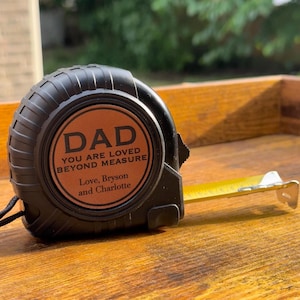 May include: Black tape measure with a copper-colored face. The face reads "DAD" above "YOU ARE LOVED BEYOND MEASURE" and "Love, Bryson and Charlotte". The tape extends with a yellow blade and a silver tip. The tape measure is on a wooden surface.