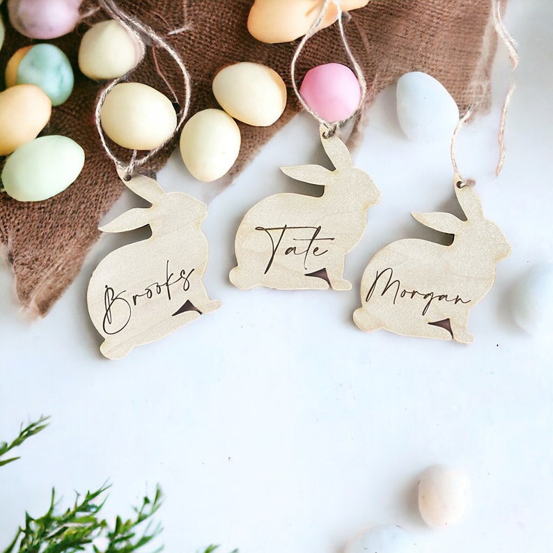 May include: Three wooden bunny-shaped ornaments with the names Brooks, Tate, and Morgan written on them. The ornaments are hanging from twine and are surrounded by pastel-colored Easter eggs.