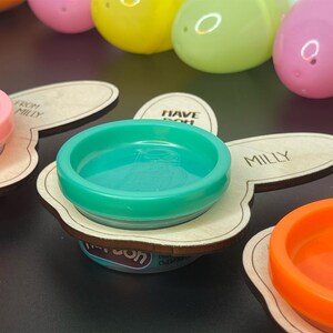 May include: Three wooden bunny-shaped holders with the text "Have fun" and "From Milly" on them. Each holder has a small container of playdough in pink, green, and orange.