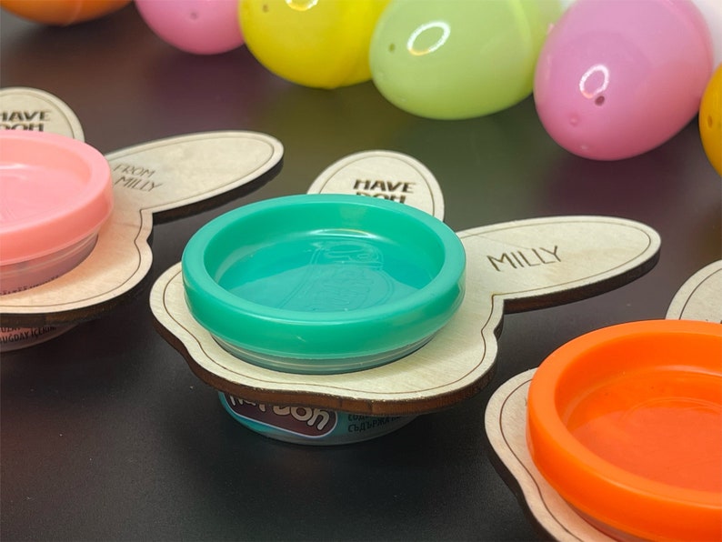 May include: Three wooden bunny-shaped holders with the text "Have fun" and "From Milly" on them. Each holder has a small container of playdough in pink, green, and orange.
