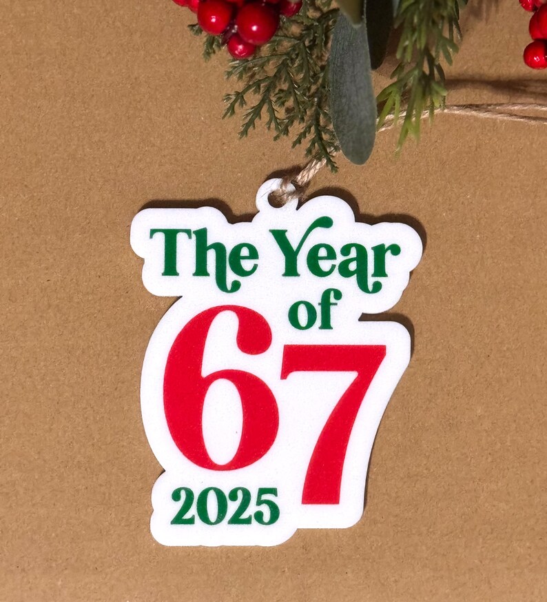 K&ouml;nnte beinhalten: Ein wei&szlig;er Weihnachtsschmuck mit dem Text "The Year of 67 2025" in Gr&uuml;n und Rot. Der Schmuck h&auml;ngt an einer nat&uuml;rlichen Schnur, vor einem braunen Hintergrund mit Zweigen und roten Beeren.