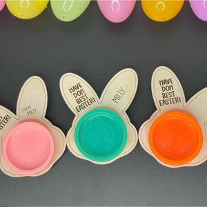 May include: Three wooden bunny-shaped Easter treat holders with the text "Have Doh Best Easter!" and "From Milly" on them. Each holder has a different color Play-Doh container inside.