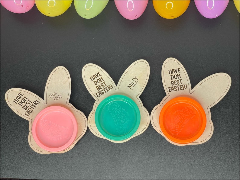 May include: Three wooden bunny-shaped Easter treat holders with the text "Have Doh Best Easter!" and "From Milly" on them. Each holder has a different color Play-Doh container inside.