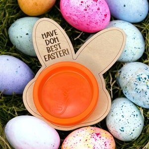 May include: A wooden bunny-shaped Easter egg holder with the text "Have Doh Best Easter!" and an orange plastic container inside. The holder is surrounded by colorful Easter eggs.