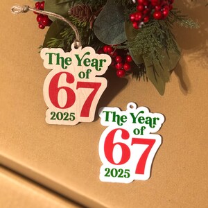 K&ouml;nnte beinhalten: Zwei Weihnachtsbaumschmuckst&uuml;cke mit dem Text "The Year of 67 2025" in Gr&uuml;n und Rot. Eines ist holzfarben, das andere wei&szlig;. Sie h&auml;ngen an Schn&uuml;ren, mit einem festlichen Arrangement aus gr&uuml;nen und roten Beeren im Hintergrund.
