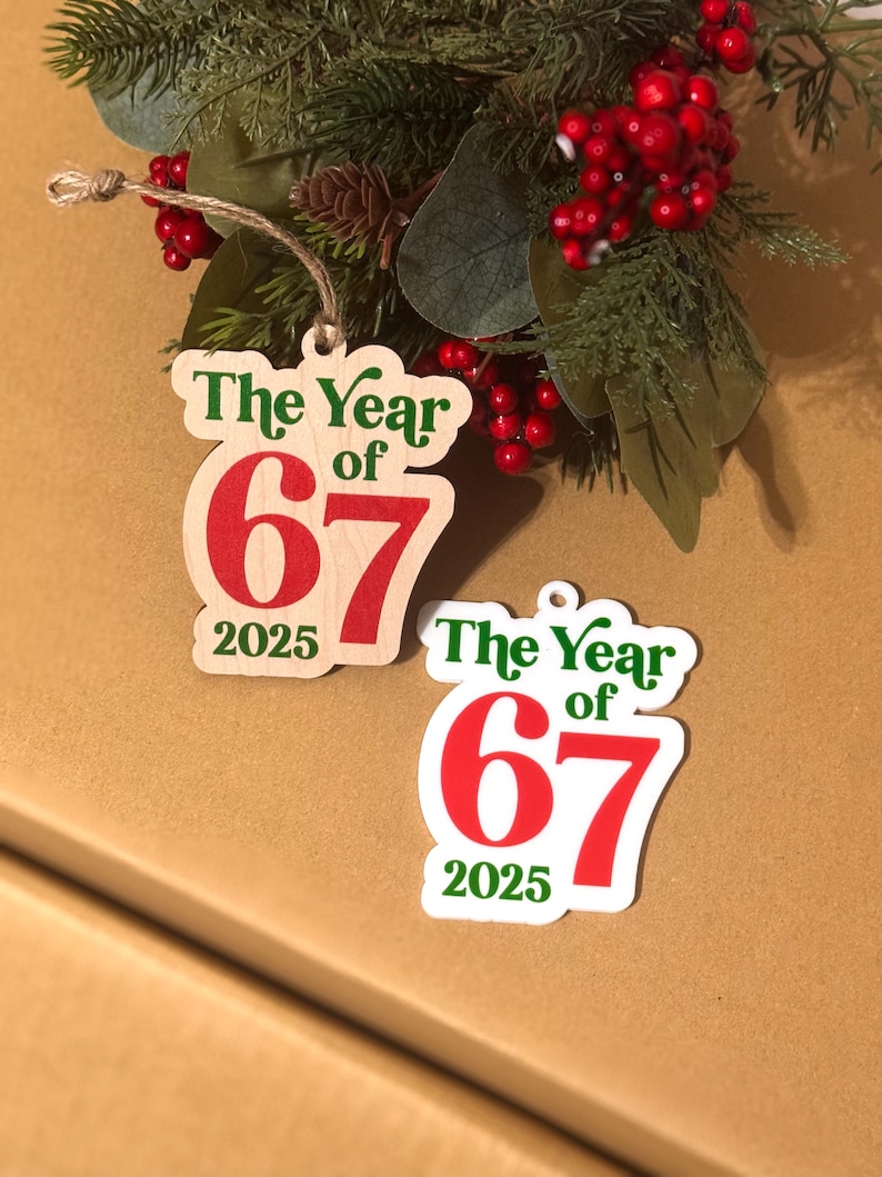 K&ouml;nnte beinhalten: Zwei Weihnachtsbaumschmuckst&uuml;cke mit dem Text "The Year of 67 2025" in Gr&uuml;n und Rot. Eines ist holzfarben, das andere wei&szlig;. Sie h&auml;ngen an Schn&uuml;ren, mit einem festlichen Arrangement aus gr&uuml;nen und roten Beeren im Hintergrund.