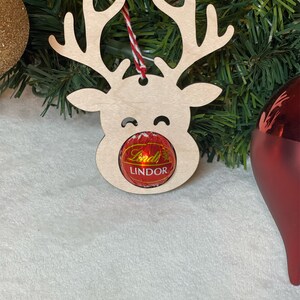 Peut inclure: Ornement de renne en bois avec une truffe au chocolat Lindor rouge pour le nez. L'ornement est suspendu &agrave; une ficelle rouge et blanche.