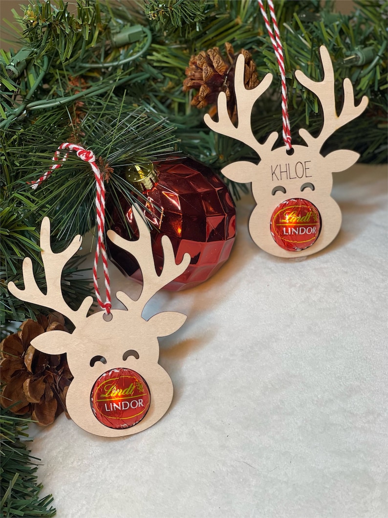 Peut inclure: Deux d&eacute;corations de rennes en bois avec une ficelle rouge et blanche suspendues &agrave; un sapin de No&euml;l. Chaque renne a un chocolat Lindor dans sa bouche. Un renne porte le nom "Khloe" &eacute;crit dessus.