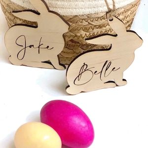May include: Two wooden bunny shaped ornaments with the names "Jake" and "Belle" engraved on them.  Two Easter eggs, one pink and one yellow, are in the foreground.