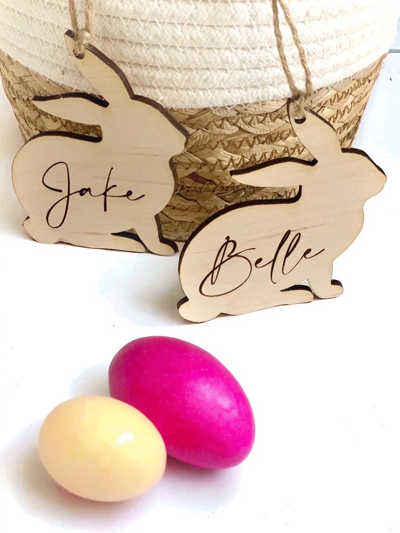 May include: Two wooden bunny shaped ornaments with the names "Jake" and "Belle" engraved on them.  Two Easter eggs, one pink and one yellow, are in the foreground.