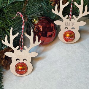 Peut inclure: Deux d&eacute;corations de rennes en bois avec de la ficelle rouge et blanche. Chaque renne a une truffe au chocolat Lindor attach&eacute;e &agrave; son nez. Un renne porte le nom "Khloe" &eacute;crit dessus.