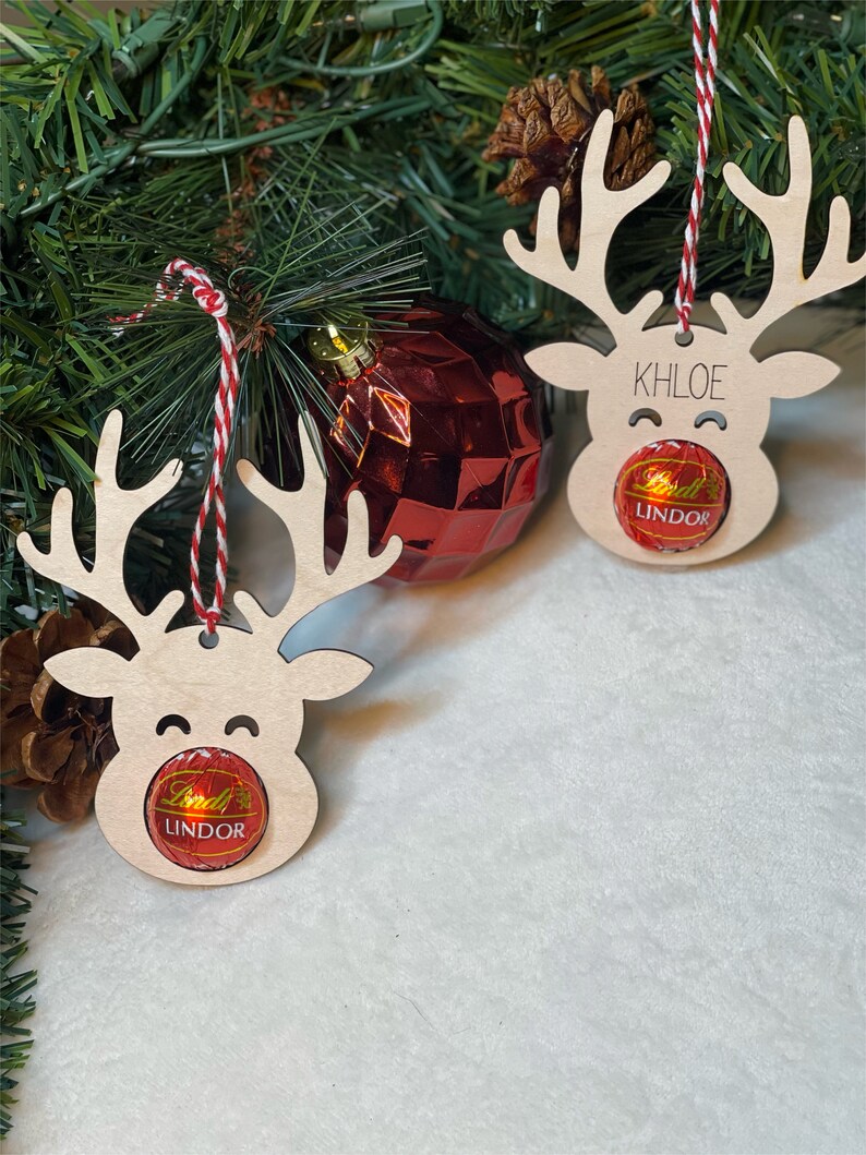 Peut inclure: Deux d&eacute;corations de rennes en bois avec de la ficelle rouge et blanche. Chaque renne a une truffe au chocolat Lindor attach&eacute;e &agrave; son nez. Un renne porte le nom "Khloe" &eacute;crit dessus.