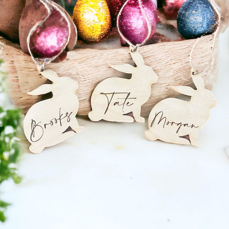 May include: Three wooden bunny-shaped ornaments with the names "Brooks", "Tate", and "Morgan" engraved on them. The ornaments are hanging from twine and are displayed against a background of colorful Easter eggs.