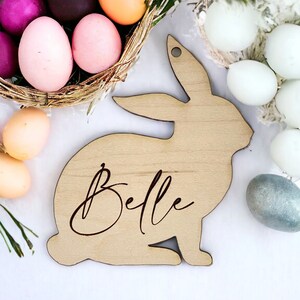 May include: A wooden bunny rabbit cutout with the name "Belle" engraved on it. The bunny is sitting on a white surface with Easter eggs in the background.