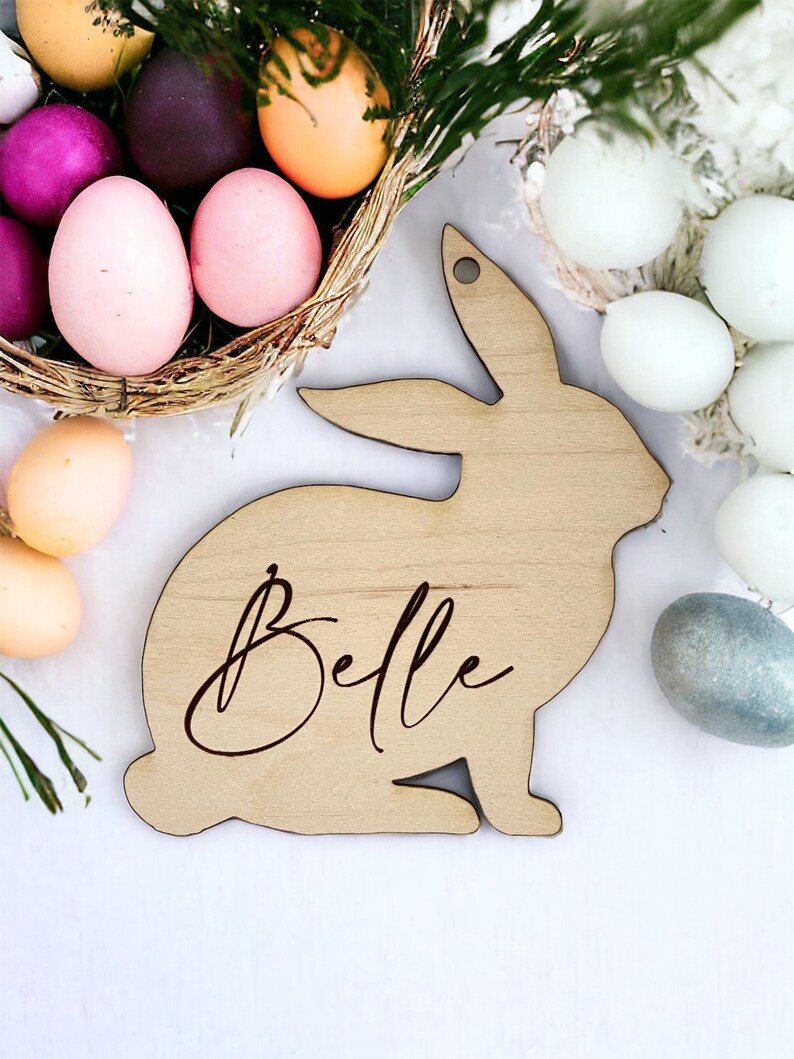 May include: A wooden bunny rabbit cutout with the name "Belle" engraved on it. The bunny is sitting on a white surface with Easter eggs in the background.