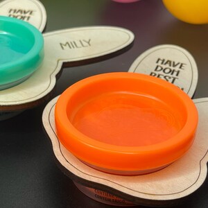 May include: Two wooden bunny-shaped trays with a small, round, orange and turquoise container on each. The containers are labeled "Milly" and "Have Doh Best".