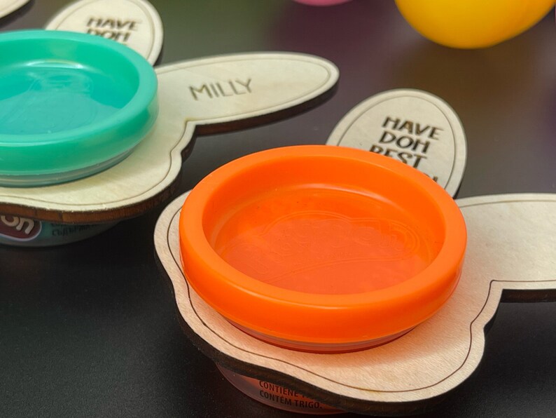 May include: Two wooden bunny-shaped trays with a small, round, orange and turquoise container on each. The containers are labeled "Milly" and "Have Doh Best".