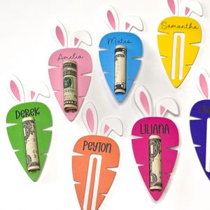 May include: Colorful carrot-shaped gift card holders with bunny ear accents. Each holder is a different color and has a name printed on it. Rolled-up dollar bills are inserted into the carrot cutouts. The names include Amelia, Mateo, Samantha, Derek, Peyton, and Liliana.