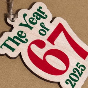 K&ouml;nnte beinhalten: Holzornament mit dem Text "The Year of 67 2025" in Gr&uuml;n und Rot. Das Ornament hat eine helle Holzmaserung und eine Schnur zum Aufh&auml;ngen.