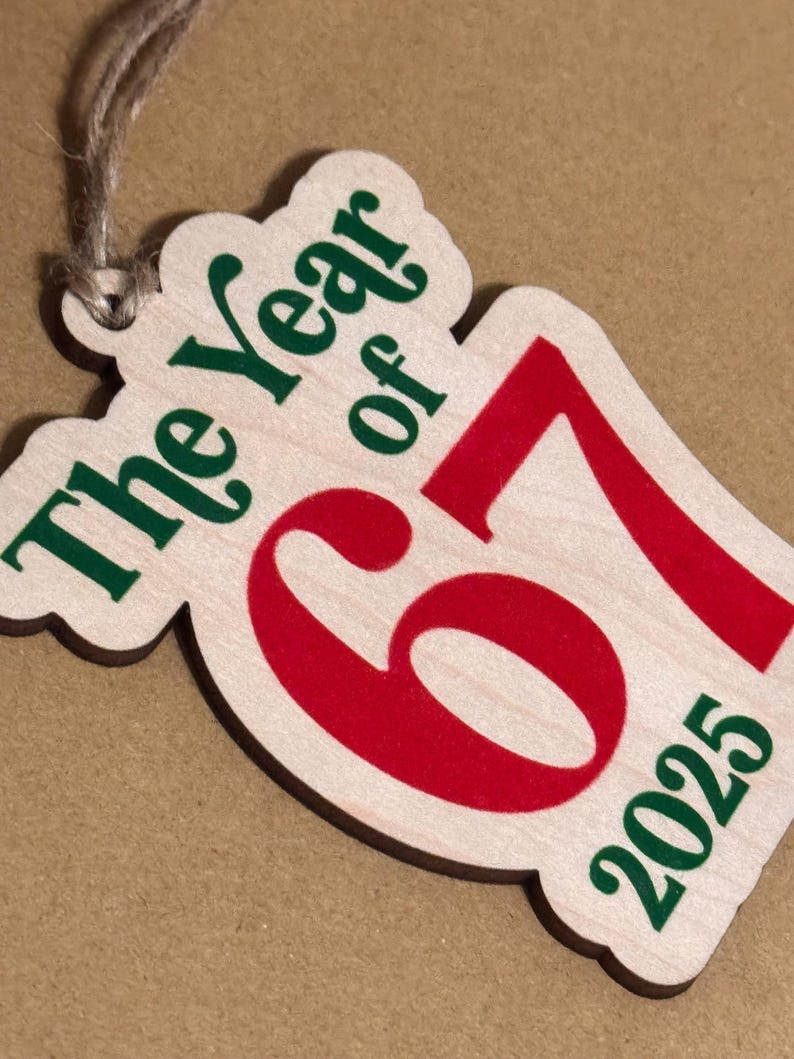 K&ouml;nnte beinhalten: Holzornament mit dem Text "The Year of 67 2025" in Gr&uuml;n und Rot. Das Ornament hat eine helle Holzmaserung und eine Schnur zum Aufh&auml;ngen.