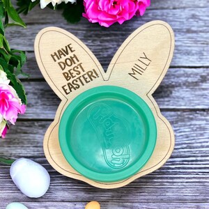 May include: A wooden bunny-shaped plate with the text "Have Doh Best Easter!" and "Milly" on it. A green plastic bowl sits on the plate. There are five pastel-colored Easter eggs on a wooden surface.