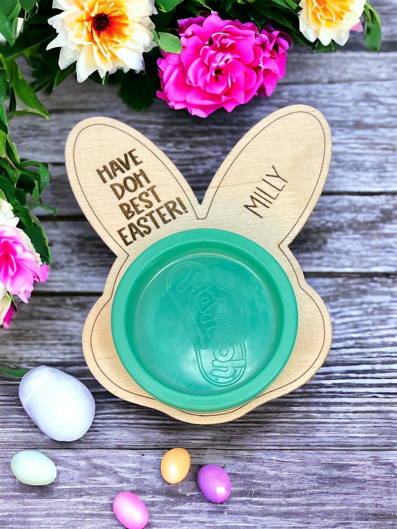 May include: A wooden bunny-shaped plate with the text "Have Doh Best Easter!" and "Milly" on it. A green plastic bowl sits on the plate. There are five pastel-colored Easter eggs on a wooden surface.