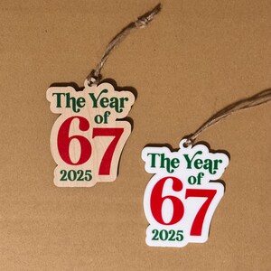 K&ouml;nnte beinhalten: Zwei Holzornamente mit dem Text "The Year of 67 2025" in Gr&uuml;n und Rot. Die Ornamente sind mit einer nat&uuml;rlichen Schnur befestigt. Ein Ornament befindet sich auf einem hellen Holzgrund, das andere auf wei&szlig;em Hintergrund.