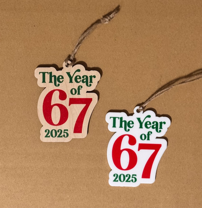 K&ouml;nnte beinhalten: Zwei Holzornamente mit dem Text "The Year of 67 2025" in Gr&uuml;n und Rot. Die Ornamente sind mit einer nat&uuml;rlichen Schnur befestigt. Ein Ornament befindet sich auf einem hellen Holzgrund, das andere auf wei&szlig;em Hintergrund.